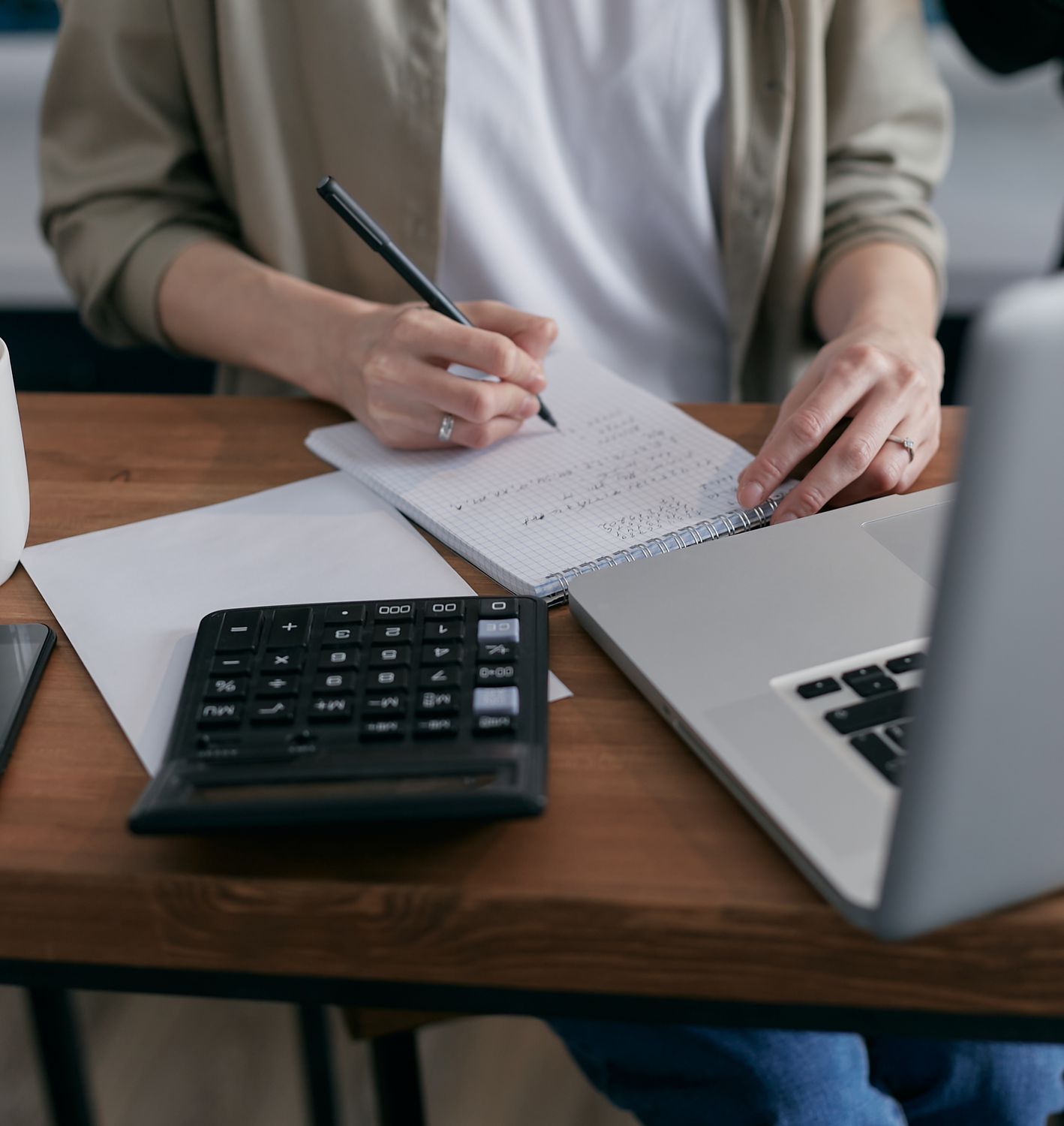 A person reviewing figures in a notebook beside a calculator and laptop